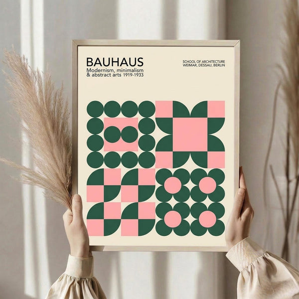May include: Person holding framed Bauhaus-inspired geometric poster with pink and dark green circles, squares, and curved shapes arranged in grid pattern. Light wood frame against neutral background with dried pampas grass visible to left. Cream colored background with minimalist typography at top.