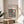 May include: Framed Bauhaus poster with geometric pattern in pink and dark green circles, squares, and curved shapes on cream background, displayed on round wooden side table next to beige upholstered chair, with plant shadow cast on white wall in bright natural lighting.