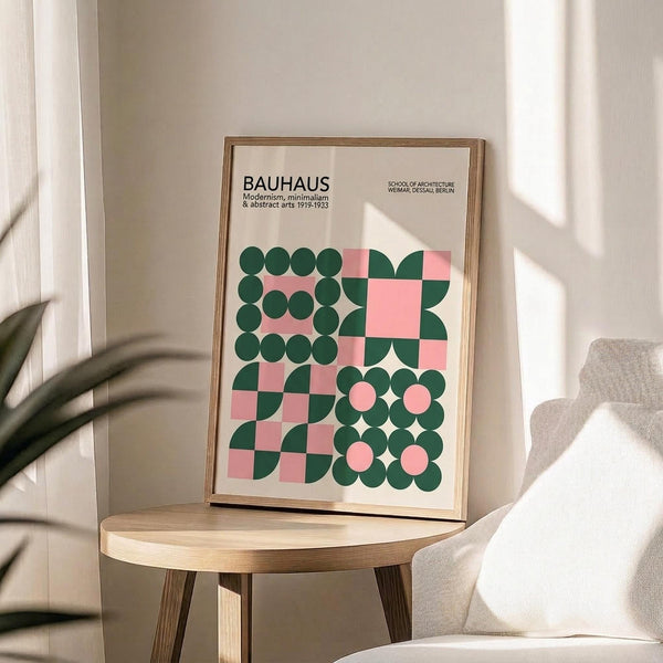May include: Framed Bauhaus poster with geometric pattern in pink and dark green circles, squares, and curved shapes on cream background, displayed on round wooden side table next to beige upholstered chair, with plant shadow cast on white wall in bright natural lighting.