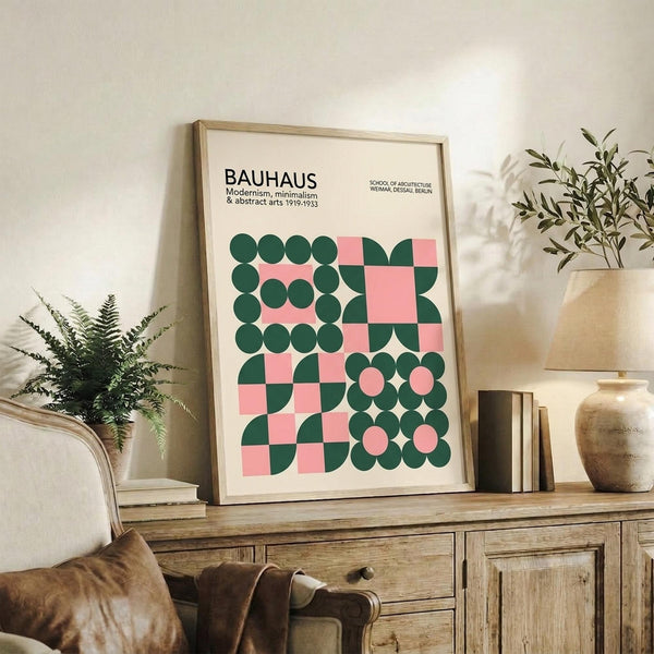 May include: Framed Bauhaus poster with geometric pattern in forest green and coral pink circles, squares, and semicircles on cream background, displayed on rustic wooden sideboard alongside potted ferns, ceramic vases, and stacked books in neutral-toned living room setting.
