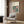 May include: Framed Bauhaus poster with pink and dark green geometric circles and squares mounted on wood paneled wall above beige upholstered armchair with patterned pillow. Modern brass floor lamp and dark round side table with open book nearby. Warm neutral interior with natural lighting from window.