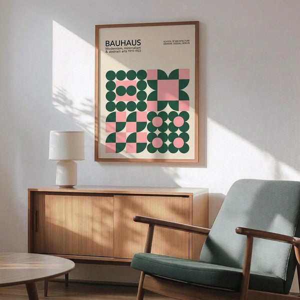 May include: Framed Bauhaus-inspired poster with geometric pattern of green circles and pink squares mounted above wooden credenza. Mid-century modern living room setting features sage green upholstered armchair with wooden arms, small round coffee table, and ceramic table lamp. Warm sunlight creates diagonal shadows across white wall, highlighting the poster's modernist design aesthetic.