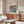 May include: Framed Bauhaus poster with geometric green and pink circles and squares displayed above a mid-century wooden credenza in a minimalist living room. White curved chair and glass coffee table visible in foreground. Warm wood tones and neutral wall colors create modern Scandinavian-inspired interior styling.