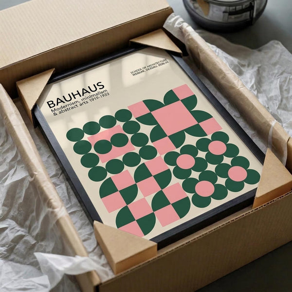 May include: Framed Bauhaus poster with geometric pattern of pink squares and green circles on cream background, shown in black frame being unpacked from cardboard shipping box with protective wrapping paper on white surface.