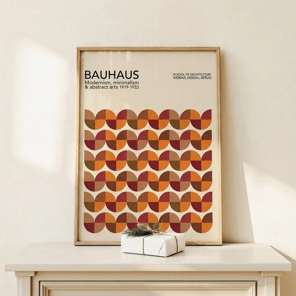 May include: Framed Bauhaus poster with geometric pattern of overlapping circles in warm autumn colors - orange, burgundy, brown, and rust tones arranged in rows. The minimalist design features text about modernism and abstract arts from 1919-1933. Natural wood frame sits on light wooden surface against cream wall, with small wrapped gift box placed in front.