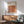 May include: Framed Bauhaus poster with geometric pattern of overlapping circles in warm orange, red, and brown tones, displayed above mid-century modern wooden credenza with white ceramic lamp. Mint green upholstered armchair with wooden frame sits nearby on wooden floor with natural sunlight casting shadows across white wall.