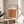 May include: A framed Bauhaus poster with geometric pattern of overlapping circles in warm autumn colors - orange, burgundy, and brown tones arranged in rows. The cream-colored print sits on a round wooden side table next to a beige upholstered chair. Natural sunlight streams through white curtains creating soft shadows, with a plant visible in the corner of this minimalist modern interior setting.