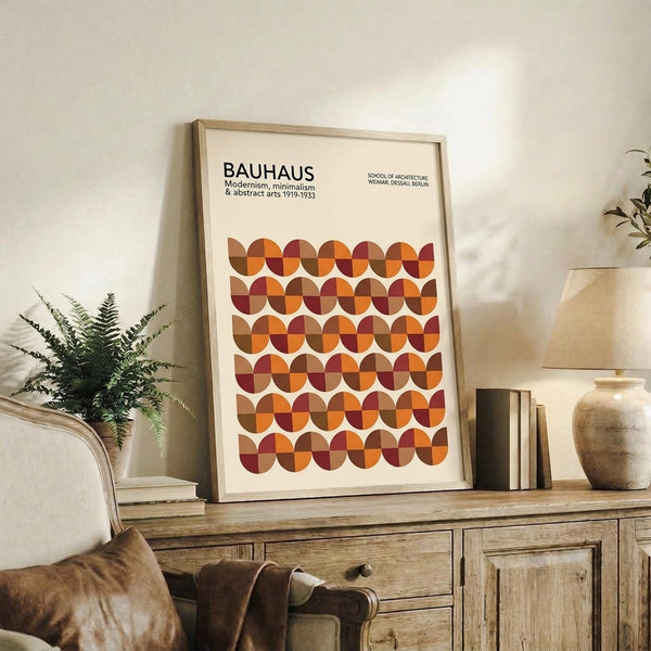 May include: Framed Bauhaus poster with geometric pattern of overlapping circles in warm autumn colors - oranges, browns, and burgundy - displayed on rustic wooden sideboard alongside fern plant, ceramic vases, and stacked books in neutral-toned living room setting