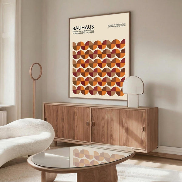 May include: Framed Bauhaus poster with geometric pattern of overlapping circles in warm orange, burgundy, and brown tones displayed above wooden sideboard in modern living room. White upholstered chair and glass-top coffee table visible. Minimalist interior with neutral walls and natural lighting creating soft shadows.