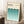May include: Framed Bauhaus poster with cream background and geometric pattern of overlapping teal semicircles in gradient shades, leaning against white wall corner. Scene shows wooden side table with white coffee cup, beige knit throw, and white area rug. Minimalist modern home decor styling with natural lighting.