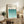 May include: Framed Bauhaus poster with teal gradient semicircle pattern displayed on rustic wooden sideboard alongside fern plant, ceramic vase, and books in warm-toned living room setting with cream upholstered chair visible