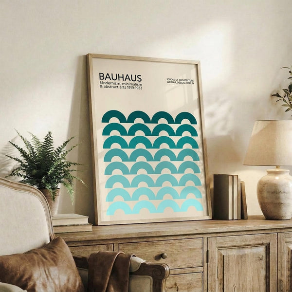 May include: Framed Bauhaus poster with teal gradient semicircle pattern displayed on rustic wooden sideboard alongside fern plant, ceramic vase, and books in warm-toned living room setting with cream upholstered chair visible