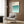 May include: Framed Bauhaus poster with teal gradient wave pattern hung on wood-paneled wall above beige upholstered armchair with geometric pillow. Modern brass floor lamp and dark side table with open book visible. Neutral interior with light wood flooring and curtained window.