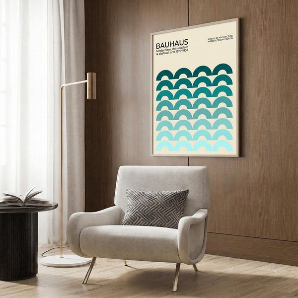May include: Framed Bauhaus poster with teal gradient wave pattern hung on wood-paneled wall above beige upholstered armchair with geometric pillow. Modern brass floor lamp and dark side table with open book visible. Neutral interior with light wood flooring and curtained window.