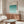 May include: Framed Bauhaus poster with teal geometric wave pattern displayed above wooden sideboard in modern living room. White sculptural chair and glass coffee table visible. Poster features graduated blue-green semicircles creating rhythmic wave design against cream background. Natural wood furniture and minimalist white table lamp complete the scene.