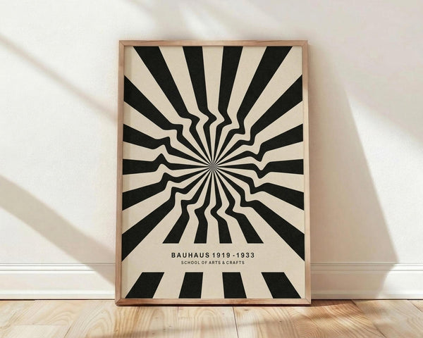 May include: A framed poster with black and cream optical illusion design featuring radiating wavy stripes that appear to curve and bend from a central point, creating a hypnotic sunburst effect. Text reads "BAUHAUS 1919-1933 SCHOOL OF ARTS & CRAFTS" at bottom. Natural wood frame leaning against white wall on light wooden floor with soft shadows from window light.