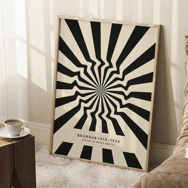 May include: Black and cream optical illusion poster with radiating wavy stripes creating a hypnotic sunburst pattern, displayed in light wood frame leaning against white wall. Small text at bottom reads "BAUHAUS 1919-1933 SCHOOL OF ARTS & CRAFTS". Scene shows cozy corner with white coffee cup on wooden stool and cream knit blanket on textured rug.