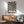 May include: Black and white optical illusion poster with radiating geometric pattern creating tunnel-like effect, displayed in thin wooden frame above mid-century modern credenza. Scene shows sage green upholstered armchair with wood arms, white ceramic table lamp, and natural sunlight casting shadows on white wall in minimalist living room setting.