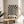 May include: Black and white optical illusion poster with radiating wavy stripes creating a hypnotic sunburst pattern, displayed in thin wooden frame on round wooden side table next to beige upholstered chair. Soft natural lighting creates shadows on neutral wall, with plant leaves visible in corner of minimalist modern room setting.