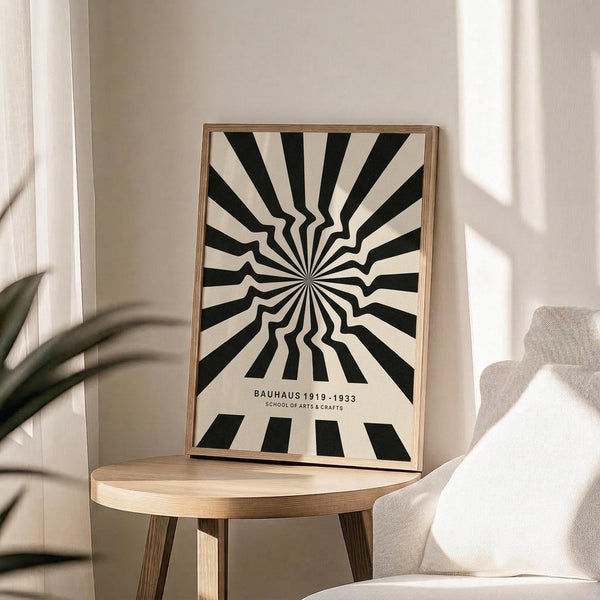 May include: Black and white optical illusion poster with radiating wavy stripes creating a hypnotic sunburst pattern, displayed in thin wooden frame on round wooden side table next to beige upholstered chair. Soft natural lighting creates shadows on neutral wall, with plant leaves visible in corner of minimalist modern room setting.