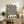 May include: Black and white optical illusion poster with radiating wavy stripes creating a hypnotic sunburst pattern, displayed in light wood frame on rustic wooden sideboard. Room scene shows cream upholstered chair, potted fern, ceramic vase, and stacked books in warm neutral living space with white walls.
