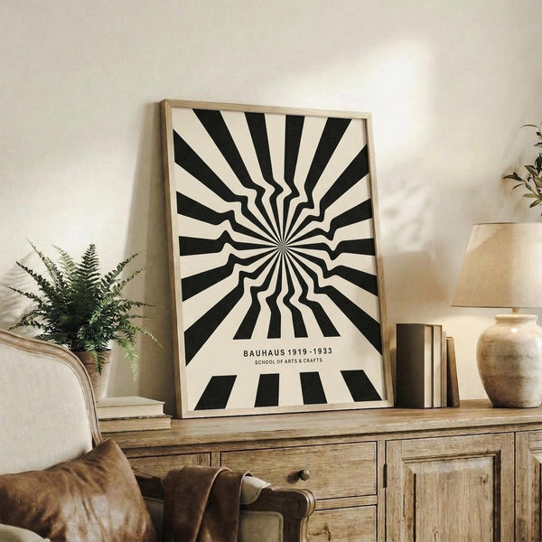 May include: Black and white optical illusion poster with radiating wavy stripes creating a hypnotic sunburst pattern, displayed in light wood frame on rustic wooden sideboard. Room scene shows cream upholstered chair, potted fern, ceramic vase, and stacked books in warm neutral living space with white walls.