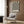 May include: Black and cream optical illusion poster with radiating stripes creating a tunnel effect, displayed on wood-paneled wall above beige upholstered armchair with geometric patterned pillow. Modern brass floor lamp and side table with open book visible. Neutral tones with natural lighting from nearby window.