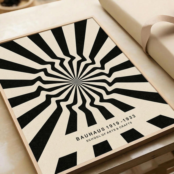 Black and white radial optical illusion design with alternating stripes radiating from center point, creating a hypnotic sunburst pattern with wavy, distorted edges. Text reads "BAUHAUS 1919-1933 SCHOOL OF ARTS & CRAFTS" in clean typography. Print shown in light wooden frame on neutral surface alongside rolled paper tubes.