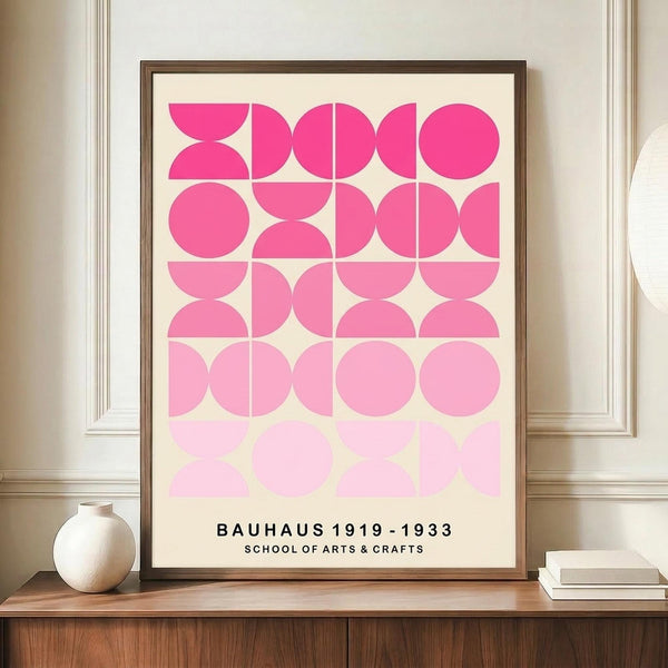 May include: Framed geometric poster with pink gradient circles and semicircles arranged in a grid pattern, displayed on wooden sideboard beside white ceramic vase and stacked books. Features "BAUHAUS 1919-1933 SCHOOL OF ARTS & CRAFTS" text at bottom. Wood frame against cream wall with decorative molding in minimalist interior setting.