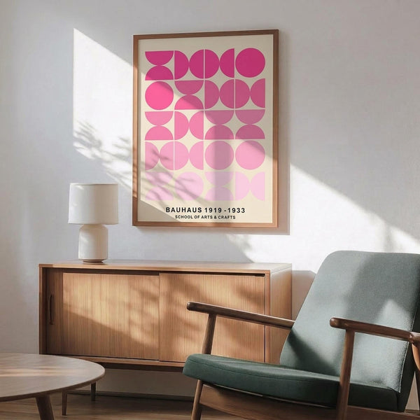 May include: Framed pink geometric art print hanging above mid-century modern wooden sideboard with white ceramic lamp. Sage green upholstered armchair with wooden frame sits nearby. Sunlight creates shadows on white wall. Print features grid pattern of pink circles and semicircles in various shades from bright fuchsia to pale pink on cream background.