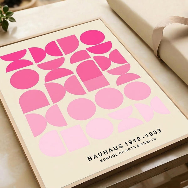May include: Pink geometric abstract art print in wooden frame displayed on neutral fabric surface next to wrapped gift with ribbon and small green plant. The artwork features overlapping circles and curved shapes in gradient pink tones from bright fuchsia to pale pink on cream background, with black text reading "BAUHAUS 1919-1933 SCHOOL OF ARTS & CRAFTS" in lower corner.
