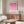 May include: Pink geometric Bauhaus poster in thin wooden frame hanging above walnut credenza in modern living room. Cream upholstered chair and glass-top coffee table visible. Poster features gradient pink circles and semi-circles in grid pattern on cream background with black text at bottom. Natural lighting creates soft shadows on white walls.