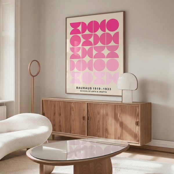 May include: Pink geometric Bauhaus poster in thin wooden frame hanging above walnut credenza in modern living room. Cream upholstered chair and glass-top coffee table visible. Poster features gradient pink circles and semi-circles in grid pattern on cream background with black text at bottom. Natural lighting creates soft shadows on white walls.
