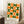 May include: Framed geometric poster with orange and green semicircles and circles arranged in a grid pattern on cream background, displayed against white wall corner. Text reads "BAUHAUS 1919-1933 SCHOOL OF ARTS & CRAFTS" at bottom. Scene shows wooden side table with white coffee cup and saucer, beige knit throw blanket, white wooden floor.
