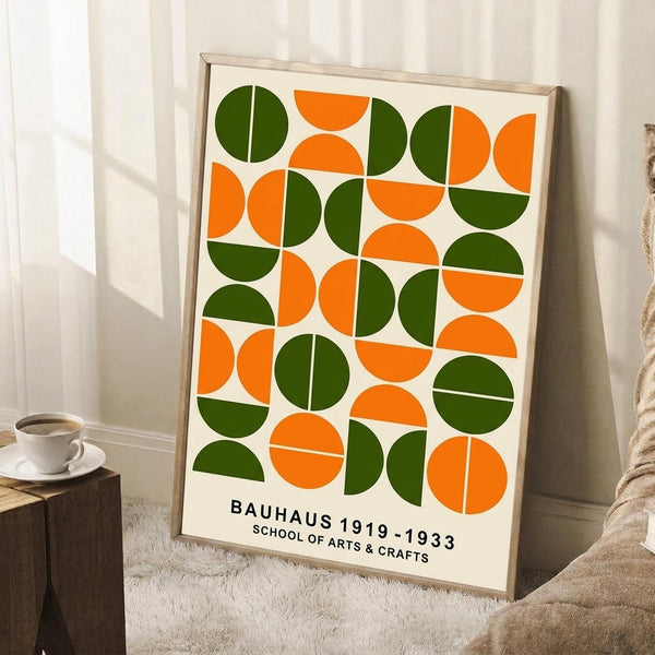 May include: Framed geometric poster with orange and green semicircles and circles arranged in a grid pattern on cream background, displayed against white wall corner. Text reads "BAUHAUS 1919-1933 SCHOOL OF ARTS & CRAFTS" at bottom. Scene shows wooden side table with white coffee cup and saucer, beige knit throw blanket, white wooden floor.