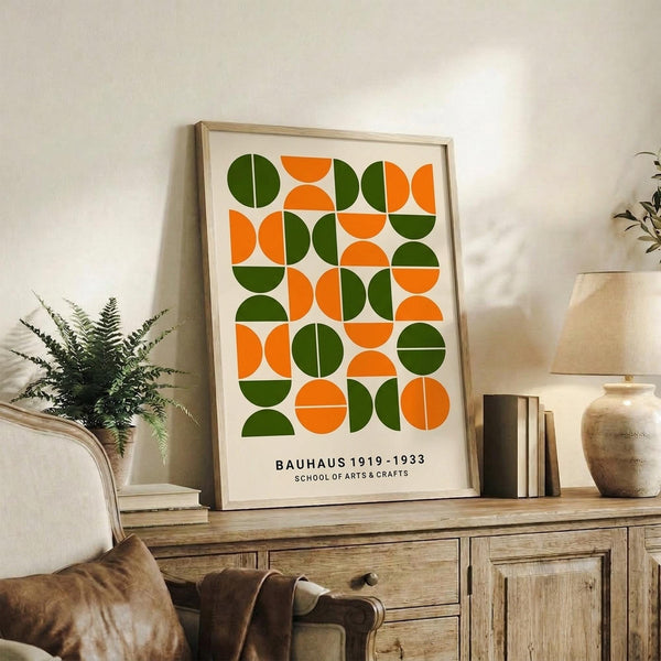 May include: Framed geometric poster with alternating green and orange circles and semicircles arranged in a grid pattern, displayed on rustic wooden sideboard alongside fern plant, ceramic vase, and stacked books in neutral-toned living room setting.
