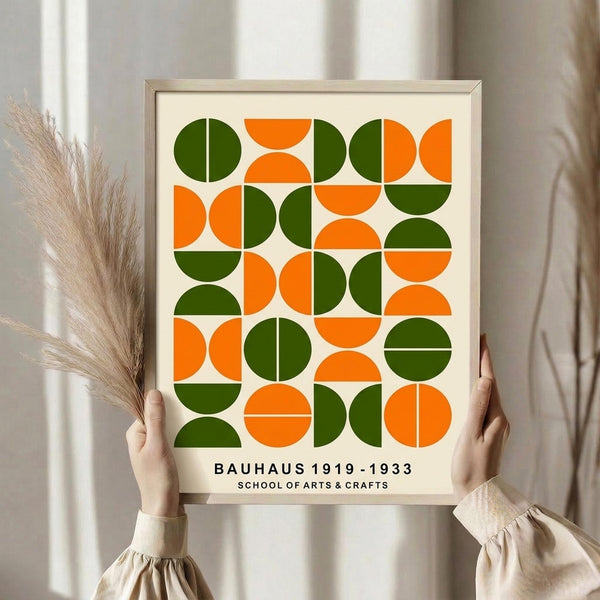 May include: Person in beige ruffled sleeves holding framed geometric art print with orange and green circles and semicircles arranged in grid pattern. Light wooden frame against cream background. Dried pampas grass visible to left. Soft natural lighting near white curtained window.