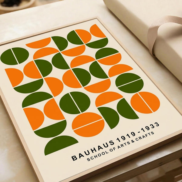 May include: Framed geometric art print featuring repeating pattern of split circles in orange and olive green on cream background. Print displays "BAUHAUS 1919-1933 SCHOOL OF ARTS & CRAFTS" text at bottom. Wooden frame sits on light wooden surface next to white sculptural objects, showing mid-century modern minimalist aesthetic with warm neutral tones.
