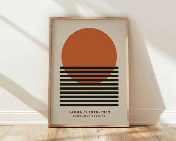 May include: Framed geometric poster featuring a large orange circle above horizontal black stripes on cream background, displayed in light wood frame leaning against white wall on wooden floor with natural lighting creating soft shadows