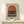 May include: Framed geometric poster with orange circle and black horizontal stripes on cream background, displayed on light wood surface next to small wrapped gift with twine and greenery sprig against white wall.