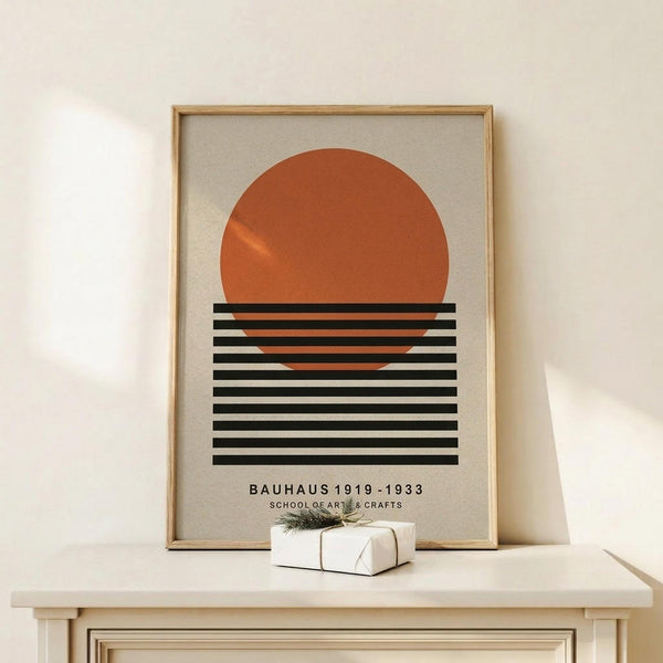 May include: Framed geometric poster with orange circle and black horizontal stripes on cream background, displayed on light wood surface next to small wrapped gift with twine and greenery sprig against white wall.