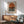 May include: Framed Bauhaus-style poster featuring an orange sunset circle above horizontal black stripes on cream background, displayed above wooden sideboard with white ceramic lamp, next to mid-century modern green upholstered chair with wooden arms in sunlit room with white walls.