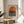 May include: Framed Bauhaus-style poster featuring orange semicircle above black horizontal stripes on cream background, displayed on round wooden side table next to beige upholstered chair. Natural sunlight streams across neutral-toned room with plant leaves visible in corner. Poster has thin wooden frame and minimalist geometric design in warm earth tones.