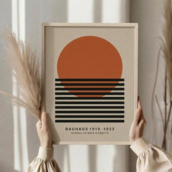May include: Person in beige outfit holding framed minimalist poster featuring orange semicircle above black horizontal lines on cream background, with dried pampas grass stems visible to the left, set against white curtained window backdrop in soft natural lighting.