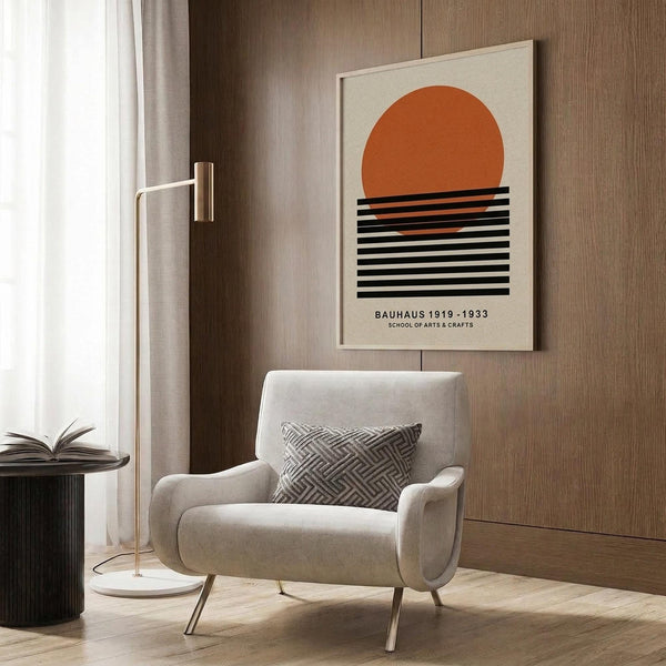 May include: Modern beige armchair with geometric patterned pillow in neutral living room setting. Framed poster on wood wall panel features orange semicircle sun above black horizontal stripes on cream background with "Bauhaus 1919-1933 School of Arts & Crafts" text below. Gold floor lamp, dark round side table with open book, light wood flooring, and white curtains complete the minimalist mid-century modern room scene.