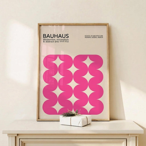 May include: Framed Bauhaus poster with pink wavy geometric pattern displayed on light wood console table against cream wall. The minimalist print features flowing curved shapes creating diamond-like negative spaces on cream background. A small wrapped gift with evergreen sprig sits beside the thin wooden frame.