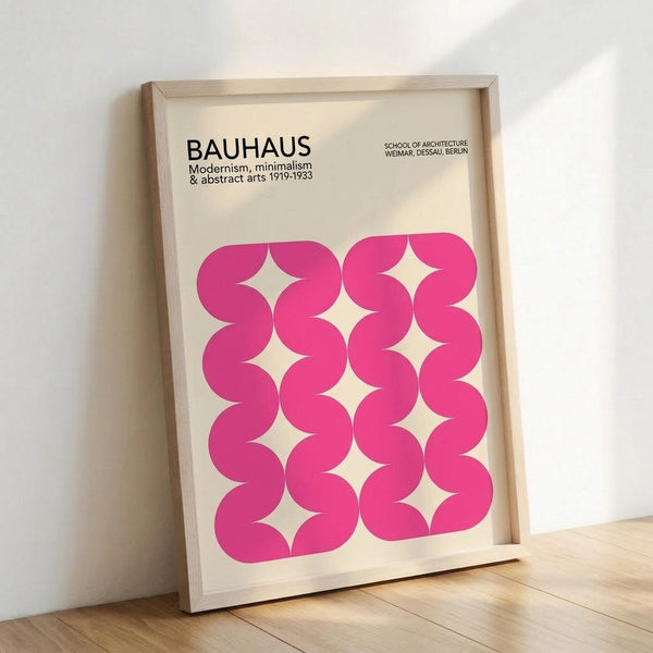 May include: A framed poster leaning against a white wall on light wooden flooring. The print features bold pink wavy shapes arranged in a grid pattern on cream background, with black text reading "BAUHAUS Modernism, minimalism & abstract arts 1919-1933" and "SCHOOL OF ARCHITECTURE WEIMAR, DESSAU, BERLIN" at the top. Natural wood frame with visible grain texture.