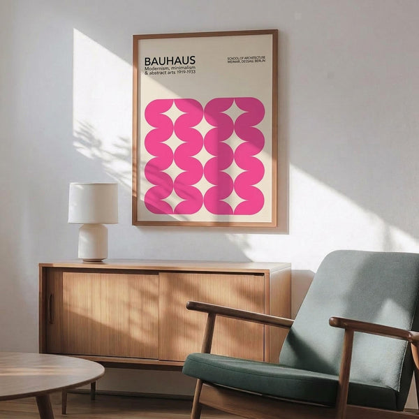 May include: Framed Bauhaus poster with pink geometric wavy pattern hanging above mid-century modern wooden credenza. Light green upholstered armchair with wooden frame sits nearby, white ceramic table lamp on credenza. Bright natural light creates diagonal shadows on white wall. Vintage-inspired interior design with warm wood tones and minimalist aesthetic.