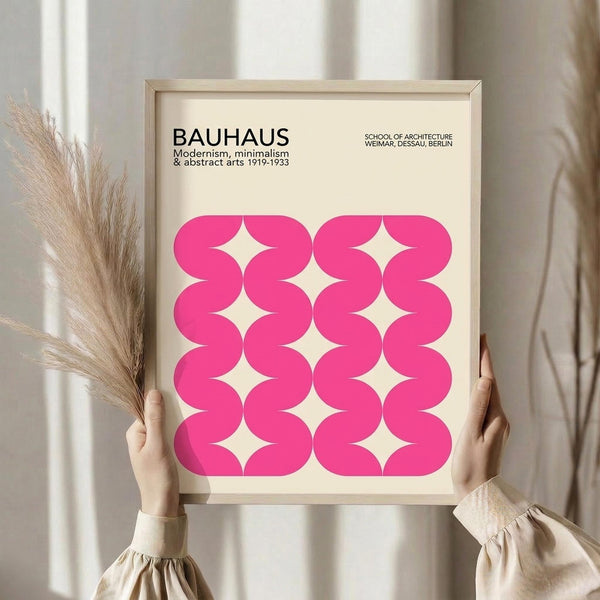 May include: Hands holding a framed poster with pink wavy geometric pattern against cream background. Frame appears light wood with clean lines. Dried pampas grass visible on left side. Person wearing beige ruffled sleeve top. Soft natural lighting creates shadows on neutral wall behind.