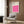 May include: Modern living room with beige armchair and geometric pillow, brass floor lamp, dark round side table with open book, and wood-paneled wall displaying framed Bauhaus poster featuring bright pink wavy geometric pattern on cream background.